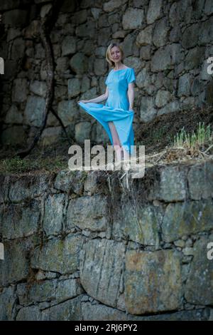 Eine Frau in einem blauen luftigen Kleid steht in mittelalterlichen Ruinen in der Nähe einer Steinmauer. Stockfoto