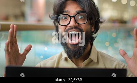Aufgeregter männlicher, bärtiger indischer Unternehmer in einer Brille, der über die Leistung eines Laptops erfreut ist. Glücklicher arabischer Geschäftsmann, der Computer nutzt Stockfoto