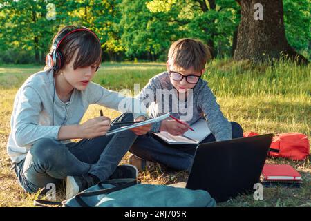 Zwei Klassenkameraden, die auf dem Boden vor dem Laptop sitzen und Hausaufgaben in Arbeitsbüchern machen. Junge in der Brille, der den Schüler im Ohr genau beobachtet Stockfoto