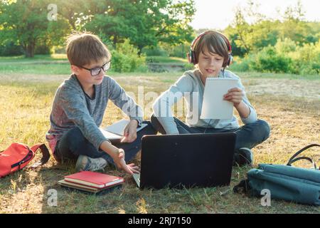 Zwei Juniorschüler, die im Park auf dem Boden sitzen und mit dem Laptop lernen. Junge in der Brille, der das Arbeitsbuch auf dem Schoß hält und dem Schüler im Kopf auf ein Gadget zeigt Stockfoto