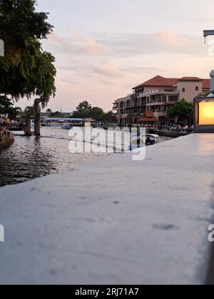 Ein kleines Boot, das an einem bewölkten Tag den malerischen Melaka River in Malaysia hinunter segelt, umgeben von wunderschönen, modernen Gebäuden Stockfoto