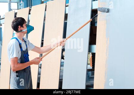 Maler in Atemschutzmaske und Overall lackiert Holzbretter mit Malerwalze auf langem Griff. Arbeiter malt Regaleinheit grau. Stockfoto