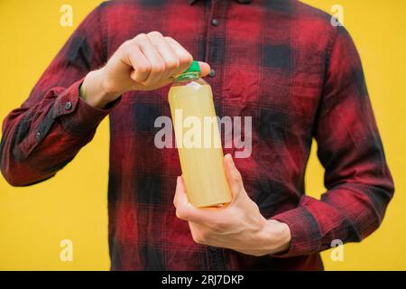 Nahaufnahme eines unbekannten Mannes in rotem Hemd, der eine orangefarbene Lemonade-Plastikflasche vor dem Hintergrund der gelben Wand im Freien hält Stockfoto