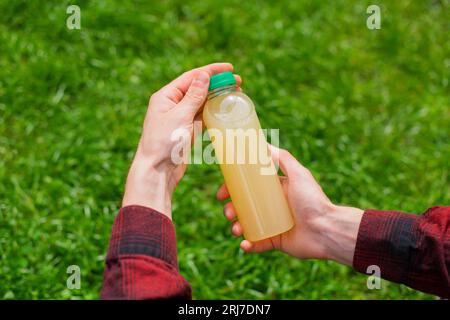 Hand eines nicht erkennbaren Mannes in einem roten Hemd, der eine gelbe Zitronenflasche in einem natürlichen grünen Grashintergrund hält, Nahaufnahme von oben Stockfoto