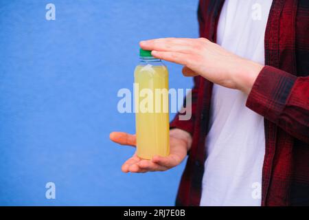 Nahaufnahme eines unerkennbaren Mannes in rotem Hemd, der Lemonade Plastikflasche vor blauem Hintergrund im Freien hält Stockfoto