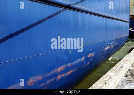 Vertikale Kratzer und Dellen am blauen Rumpf des im Hafen verankerten Containerschiffs. Auf dem Farb- und Kopierraum befinden sich einige rostige Flecken. Stockfoto