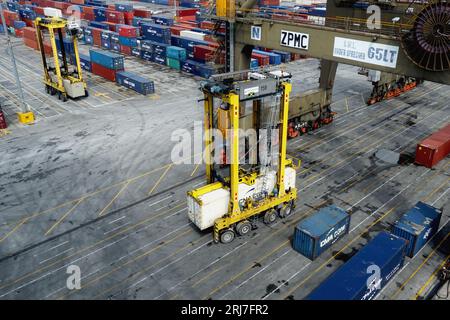 Ein gelber Radträger, beladen mit weißem Kühlcontainer im Kingston Freeport Terminal, der unter dem Portalkran vorbeifährt. Stockfoto