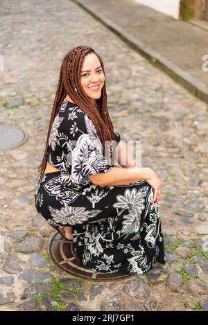 Eine Frau hockte auf einer Kopfsteinpflasterstraße. Haargeflechte und afrikanisches Kleid. Pelourinho, Brasilien. Stockfoto