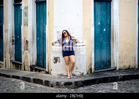 Eine hübsche Frau, die eine Sonnenbrille mit geflochtenen Haaren trägt, lehnt sich gegen die Fassade eines alten Hauses. Pelourinho, Brasilien. Stockfoto