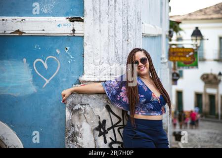 Eine hübsche Frau, die eine Sonnenbrille mit geflochtenen Haaren trägt, lehnt sich gegen die Fassade eines alten Hauses. Pelourinho, Brasilien. Stockfoto