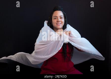 Foto einer schönen Frau mit Zöpfen im Haar in roter Kleidung, die in ein weißes Tuch gewickelt ist. Studioporträt mit schwarzem Hintergrund. Stockfoto