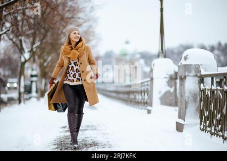 Fröhliche stilvolle Frau mittleren Alters mit braunem Hut und Schal im Kamelmantel mit Handschuhen und Einkaufstaschen im Winter draußen in der Stadt. Stockfoto