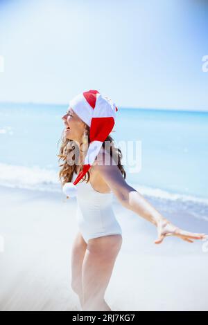 Lächelnde, stilvolle Frau mittleren Alters in weißer Strandkleidung mit gestreifter weihnachtsmütze, die Spaß am Strand hat. Stockfoto