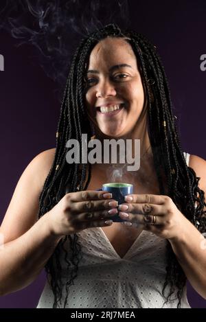 Schöne Frau mit Zöpfen im Haar, die eine Tasse Kaffee hält. Ich trage weiße Kleidung. Isoliert auf dunklem Fliederhintergrund. Positiv pro Stockfoto