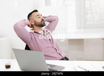 Glücklicher, ruhiger, selbstbewusster kaukasischer Reifer im Hemd mit geschlossenen Augen sitzt am Tisch mit Laptop und Ruhepausen Stockfoto