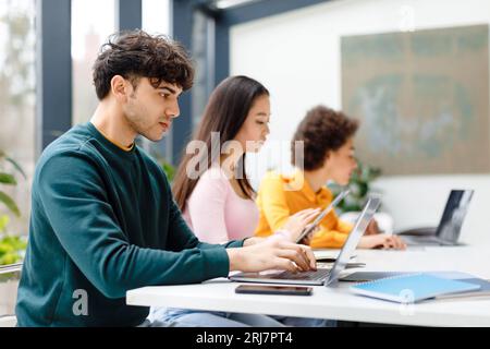 Multirassische männliche und weibliche Studenten, die für Tests studieren, Hausaufgaben mit Gadgets machen, fokussierter Kerl mit Laptop Stockfoto