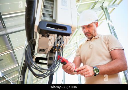 Unteransicht des Arbeiters, der die Arbeit des Solar-Wechselrichters prüft. Messen der Ausgangsspannung mit Stromzangen. Einstellen des Wechselrichters an der Rückseite des PV-Panels. Stockfoto