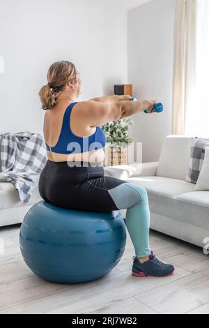 Übergewichtige Dame, die im Wohnzimmer Sport treibt. Er sitzt auf einem Fitnessball und hält seine Hände mit Kurzhanteln vor sich. Stockfoto