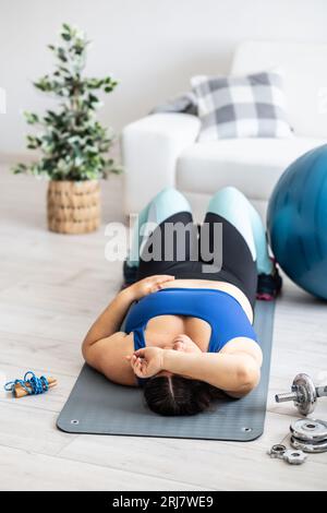 Eine müde Frau in Übergröße liegt auf einer Matte im Wohnzimmer, Sportausrüstung um sie herum. Stockfoto