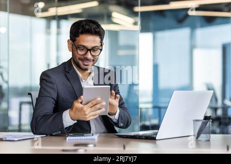 Junger erfolgreicher hispanischer Geschäftsmann am Arbeitsplatz, der App auf Tablet-Computer verwendet, lächelnder Mann im Bürogebäude, mit Laptop arbeitet, Business-Anzug trägt, zufrieden mit Erfolg. Stockfoto