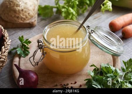 Hühnerbrühe oder Suppe in einem Glas auf einem Tisch, mit frischem Gemüse und Kräutern Stockfoto