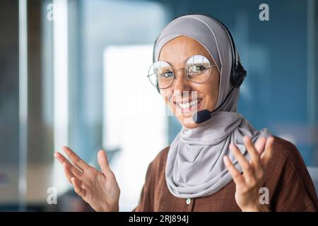 Porträt einer jungen muslimischen Lehrerin in Hijab und Headset, die Online-Schulungen durchführt. Er lächelt die Kamera an und bewegt mit den Händen. Nahaufnahme. Stockfoto