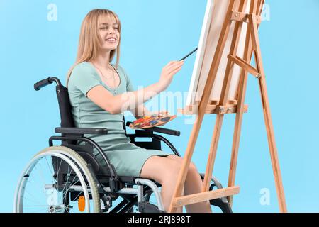 Junge Frau im Rollstuhlmalerei auf Staffelei vor blauem Hintergrund Stockfoto