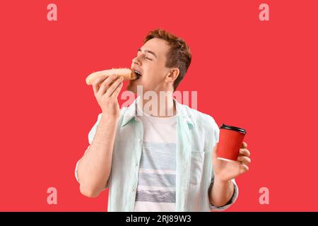 Junger Mann mit leckerem Hot Dog und einer Tasse Kaffee auf rotem Hintergrund Stockfoto