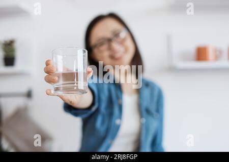 Konzentrieren Sie sich auf Glas, das halb voll mit sauberer Flüssigkeit ist und von einer glücklichen asiatischen Dame in Freizeitkleidung erreicht wird. Eine fröhliche Hausfrau, die ermutigt, sich zu wohlfühlen Stockfoto