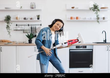 Asiatische attraktive Frau, die Musik über kabellose Kopfhörer hört und einen Staubsauger wie eine Gitarre hält. Eine energiegeladene Frau, die sich sie wie ein Mitglied von Ro vorstellt Stockfoto