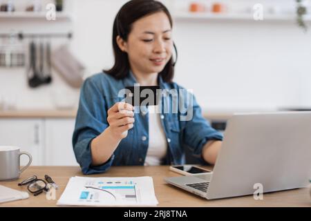 Schöne asiatische Frau mit bob-Frisur, die Kreditkarte hält und Daten in einen persönlichen Laptop eingibt. Aufmerksame Frauen, die sich mit Online-Arbeit beschäftigen Stockfoto