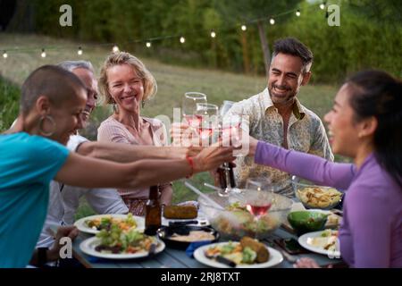 Gruppe von Freunden mittleren Alters, die gemeinsam auf der Terrasse eines Hauses oder Restaurants speisen. Glückliche Menschen essen und trinken Toast an einem Tisch im Freien Stockfoto