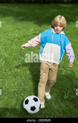 Vertikales Porträt des aktiven Tween Boy, der im Sonnenlicht auf grünem Gras Fußball spielt Stockfoto