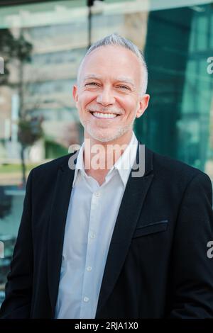 Vertikales Porträt eines Reifen erwachsenen Geschäftsmannes mit grauem Haar und Anzug, der lächelt und die Kamera mit erfolgreicher Einstellung betrachtet. Glücklicher Unternehmensanwalt mit weißen perfekten Zähnen, die am Arbeitsplatz stehen. Hochwertige Fotos Stockfoto