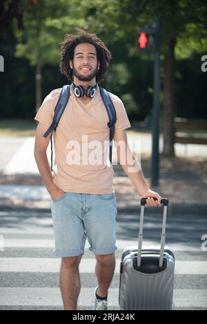 Junger gutaussehender Mann, der mit seinem Koffer die Straße überquert Stockfoto