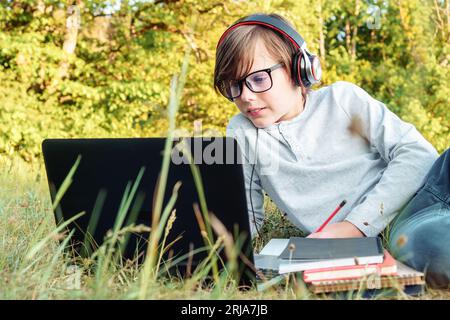 Konzentrierte Juniorschüler in Brille und Ohrhörern auf den Kopf aufmerksam die Übungen auf dem Laptop beobachten, Schreibaufgaben im Notebook schreiben, während sie auf dem Rasen in Gleichheit liegen Stockfoto