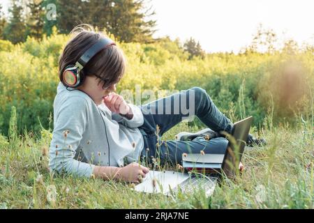 Leidenschaftlich für Studienkind in Brille, Kopfhörer, die im Park auf Gras liegen, aufmerksames Betrachten von Aufgaben auf dem Laptop. Die Pupille hält die Hand nah an der Gesichtsstudye Stockfoto