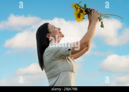 Ein Symbol des Friedens. Ein schönes und glückliches weißes Mädchen steht am blauen Himmel und hält einen Blumenstrauß mit gelben Sonnenblumen in der Hand Stockfoto