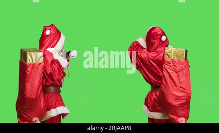 Pünktlich überprüft der weihnachtsmann die Zeit auf der Armbanduhr, während er einen roten Sack mit Geschenken trägt und ein festliches Kostüm auf dem Greenscreen trägt. Saint Nick wartet mit einer großen Tüte mit Geschenken und Spielzeug. Stockfoto