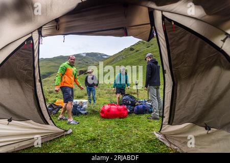 Der offene Eingang zu einem Zelt umrahmt den Campingplatz einer Trekkinggruppe auf einer Bergwiese nahe Dartlo in der Gemeinde Achmeta, Region Kachetien, Georgien. Vier Männer stehen zwischen Rucksäcken, Taschen und Kisten. Links, in einer orange-grünen Jacke, steht der Wanderführer Giorgi Chachua. Vor ihm liegt eine rote Reisetasche. Rechts steht ein Mann mit einer Schirmmütze und einer dunklen Kapuzenjacke. Im Hintergrund steigen grüne Hänge an. Links steht die Ruine eines Turms. Die Trekkingtour mit Packpferden hat fast 120 Kilometer und mehr als 5.000 Meter Höhenmeter zurückgelegt. T Stockfoto