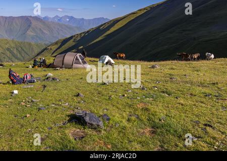 Zwei Zelte stehen auf der Bergwiese am Morgen im Lager unter dem Atsunta-Pass auf der Khevsur-Seite in der Munizipalität Duscheti, Region Mtskheta-Mtianeti, Georgien. Links vom großen braunen Zelt sitzen vier Personen an einem Tisch beim Frühstück. Sattlereien liegen neben dem kleineren weißen Zelt. Packpferde stehen auf der Wiese hinter ihnen, darunter ein graues Pferd rechts. Rucksäcke und Steine liegen im Gras im Vordergrund. Bergketten reihen sich im Hintergrund links. Der Hang rechts liegt noch im Schatten. Wanderer und Packpferde verbrachten die Nacht auf einer Höhe von etwa 3, Stockfoto