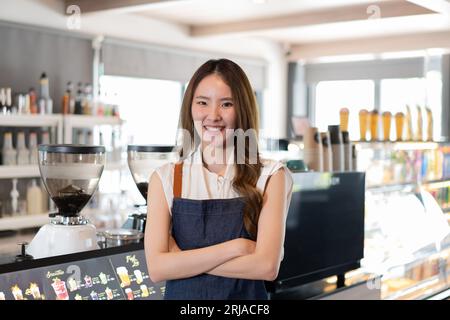 Junge Erwachsene asiatische Geschäftsfrau kreuzte vor dem Café-Restaurant ihre Arme und lächelte. Glücklicher und fröhlicher Barista im Coffee Shop, der die Arme gekreuzt hat. Stockfoto