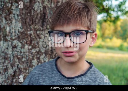 Porträt eines liebenswerten Jungen mit blauen Augen, in Brille mit grauem Longsleeve vor Baum im Park stehend. Jugendschüler mit Maulwurf über Augenbraue en Stockfoto