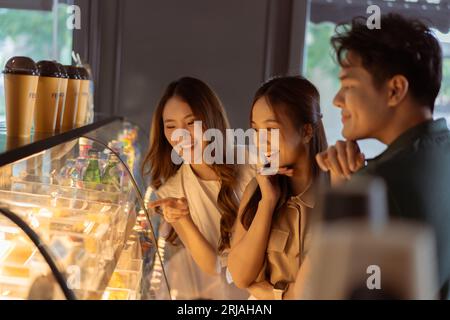 Junge asiatische Frauen, die sich Brot und Desserts ansehen, während sie aus dem Fenster der Bäckerei stehen, zeigen mit männlichen und weiblichen Freunden. Gruppe asiatischer Teenager, die im Café zeigen und bestellen Stockfoto