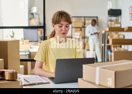 Junge Frau, die auf der Tastatur ihres Laptops schreibt, während ihr afroamerikanischer Kollege Tablet an Regal mit Kartons mit bestellten Waren benutzt Stockfoto