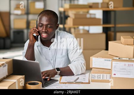 Junger afroamerikanischer Mann in weißem Hemd sitzt am Arbeitsplatz im Lager oder auf dem Markt und spricht mit dem Kunden über das Mobiltelefon Stockfoto