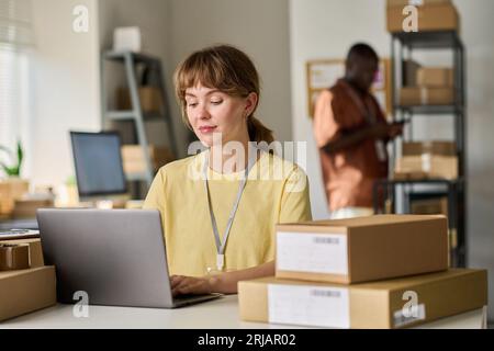 Junge hübsche Lagerarbeiterin, die Laptop gegen einen afroamerikanischen Kollegen verwendet, während sie neue Online-Bestellungen von Kunden entgegennimmt Stockfoto