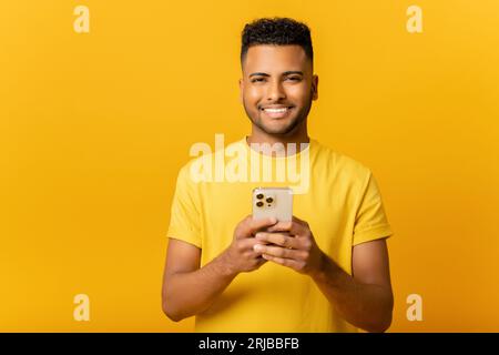 Porträt eines fröhlichen jungen indischen Mannes, der eine Nachricht auf dem Handy auf gelbem Hintergrund sendet. Glücklicher arabischer Mann, der Smartphone nutzt, online chattet und in sozialen Netzwerken Nachrichten sendet Stockfoto