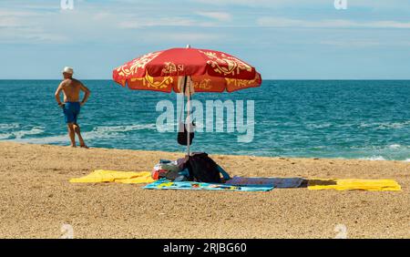 Der Mann steht am Meer in der Nähe eines Sonnenschirms, der Habseligkeiten in Nazare, Portugal, schützt Stockfoto