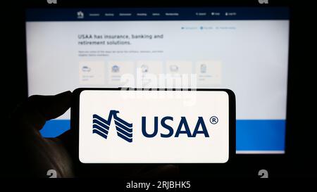 Person holding cellphone with logo of United Services Automobile Association (USAA) on screen in front of webpage. Focus on phone display. Stock Photo